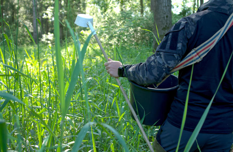 Ein KABS-Mitarbeiter steht in einer überschwemmten Fläche im Auwald und trägt einen Eimer mit Bti und einen Schöpfer.