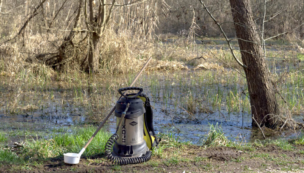 Eine Rückenspritze und ein Schöpfer sind vor einer Brutstätte im Sumpfwald abgestellt.