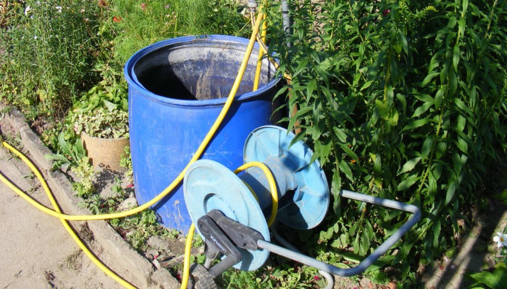 Eine offene Regentonne steht neben einer abgerollten Wasserschlauchtrommel.