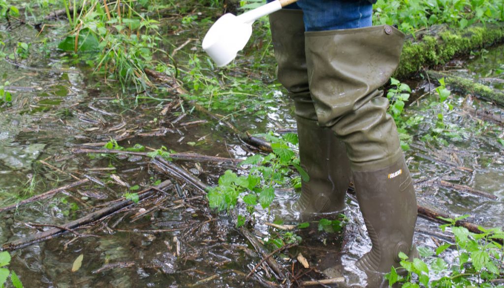 Ein KABS-Mitarbeiter steht in einer wasserführenden Senke. Nahaufnahme der Gummistiefel und des Dippers.